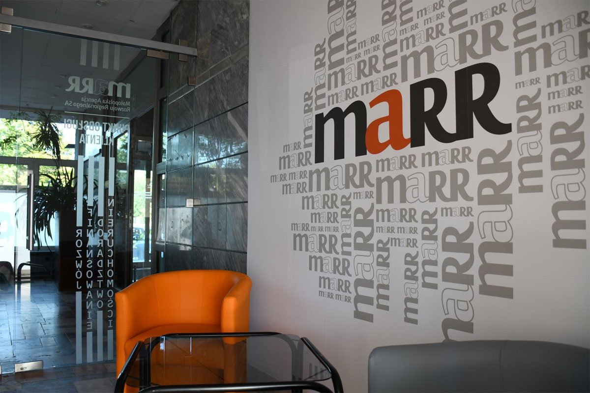 Wnętrze nowoczesnego biura – jasna przestrzeń z przeszklonym korytarzem. Na ścianie widoczne duże logo MARR