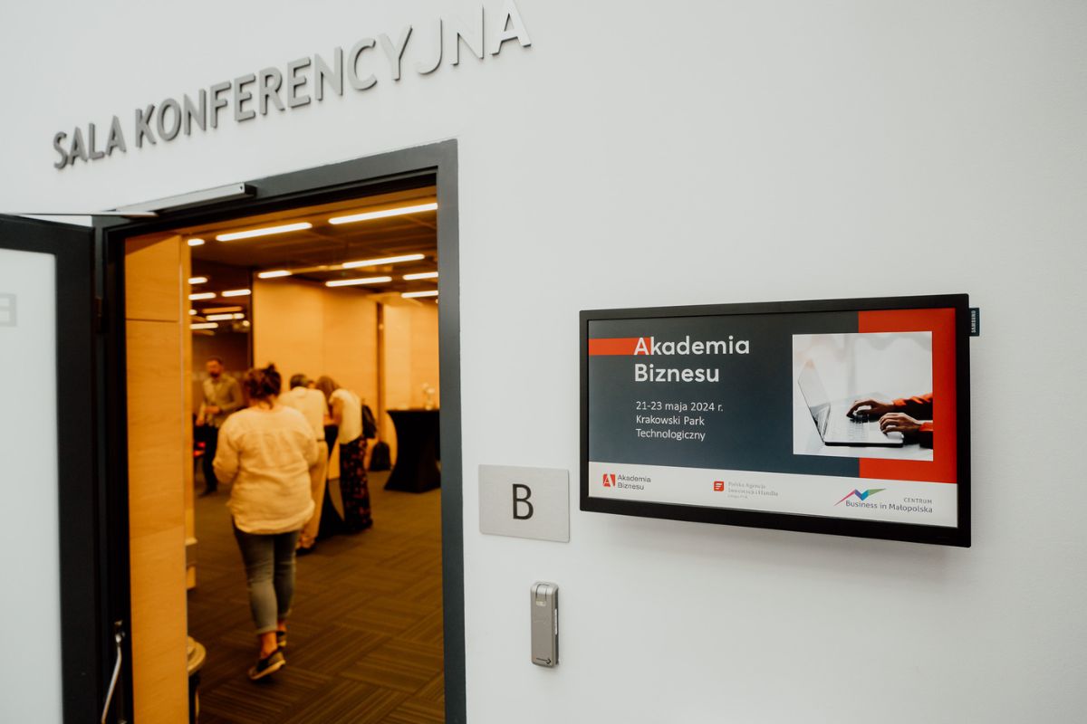Monitor z wyświetloną nazwą wydarzenia obok drzwi prowadzących na salę konferencyjną