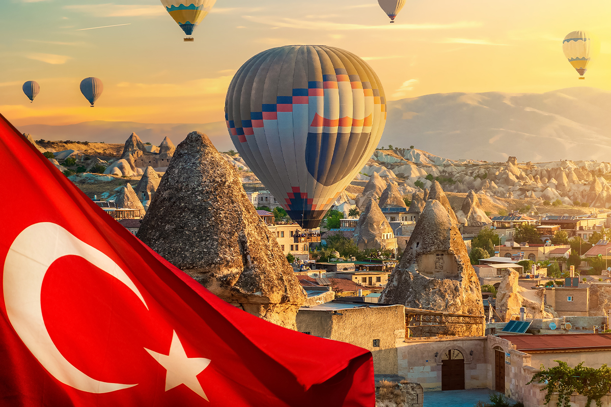 Na pierwszym planie czerwona flaga Turcji, na drugim planie starożytne kamienne budowle o stożkowym kształcie i kolorowe balony unoszące się w powietrzu