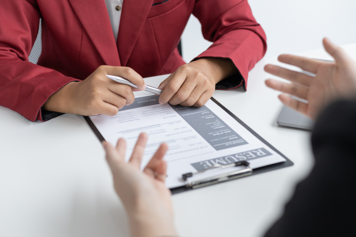 dwie osoby siedzące po przeciwnych stronach biurka prowadzące rozmowę kwalifikacyjną, na biurku CV