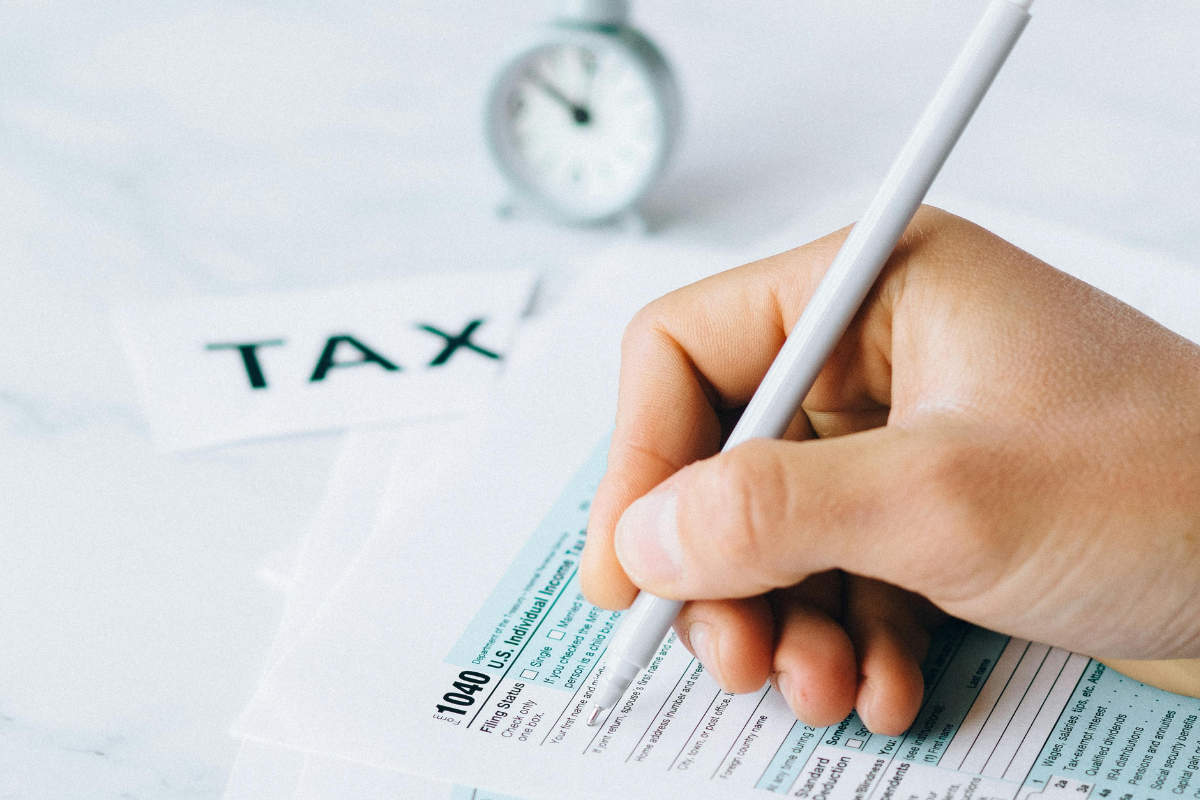 kartka formularza a nad nią dłoń trzymająca długopis, w tle napis tax oraz zegarek