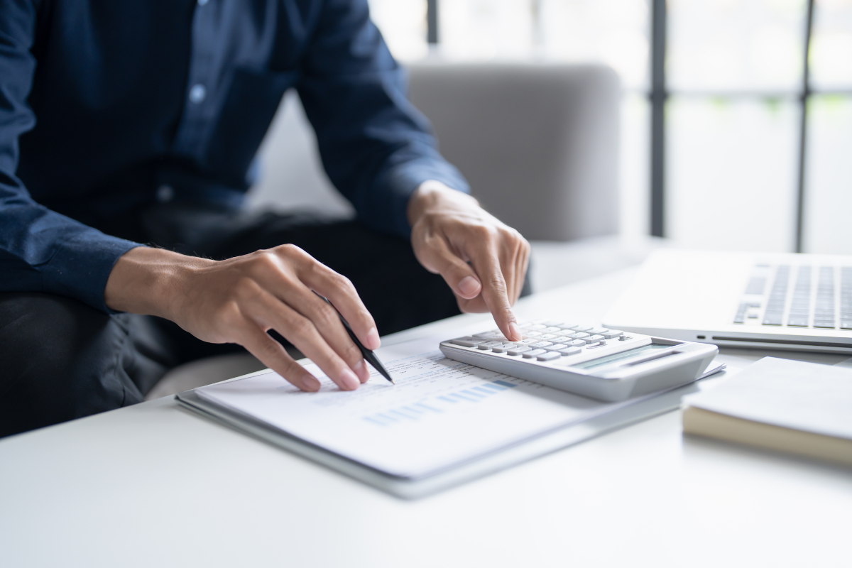na biurku otwarty laptop, dokumenty a na nich kalkulator, przy biurku siedzi osoba trzymająca w prawej dłoni długopis nad dokumentem z palcem wskazującym lewej dłoni na kalkulatorze